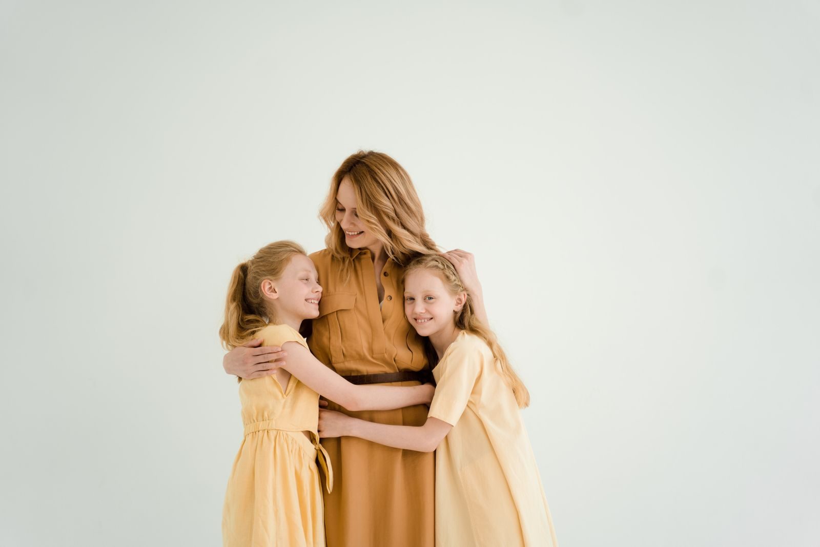 A mother lovingly embraces her two daughters against a soft, light background, symbolizing family love and togetherness.