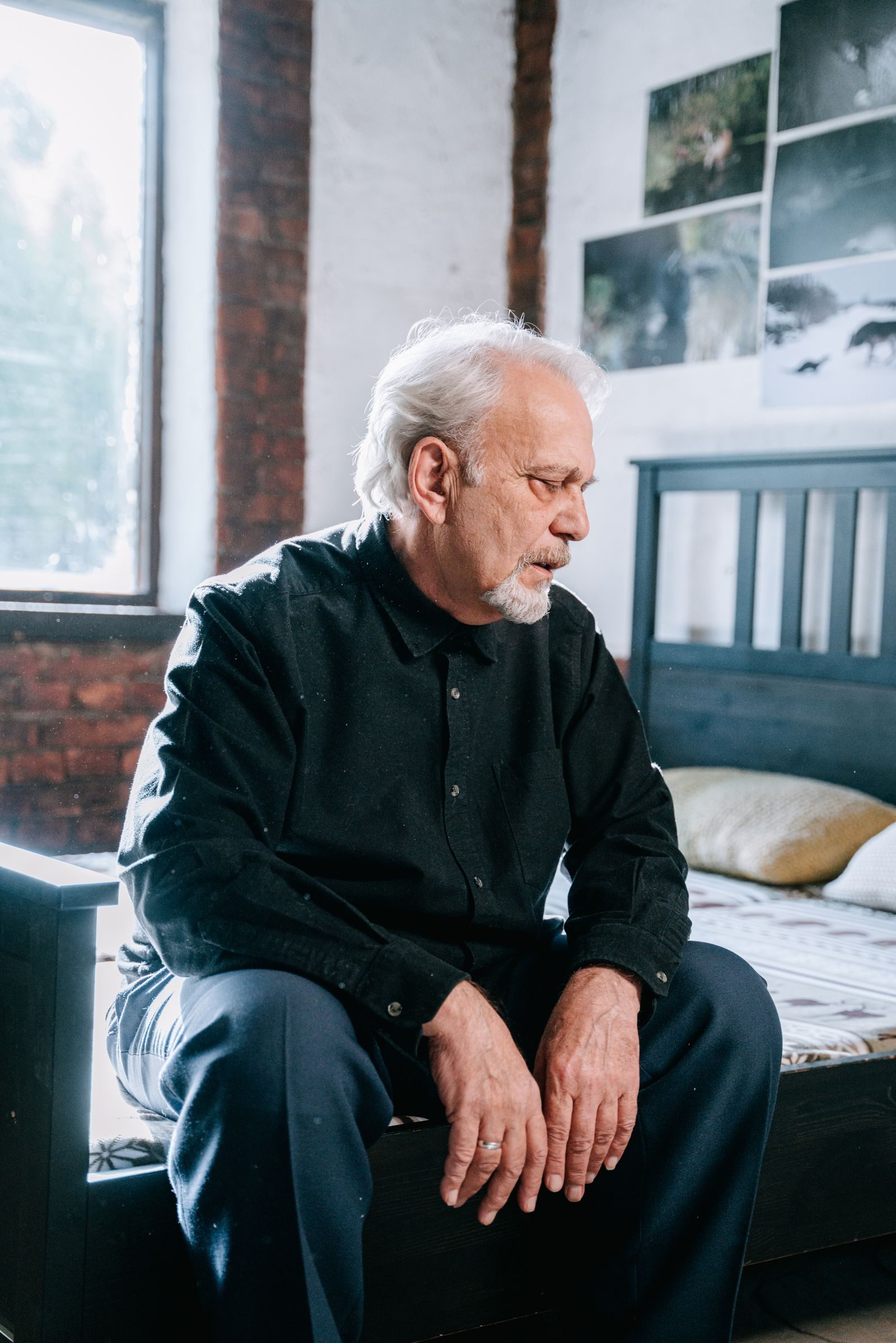 An elderly man in a black shirt sits pensively on a bed in a bright bedroom.