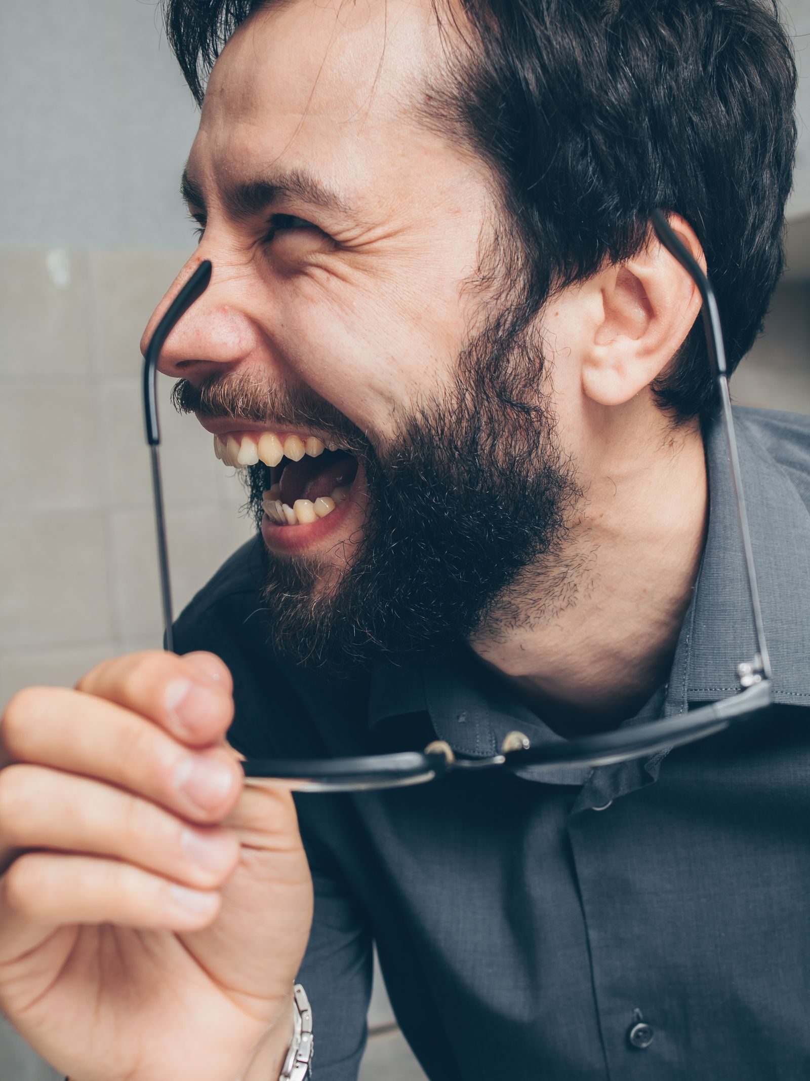 Close-up of a bearded man laughing joyfully, holding eyeglasses. Energetic and happy expression.