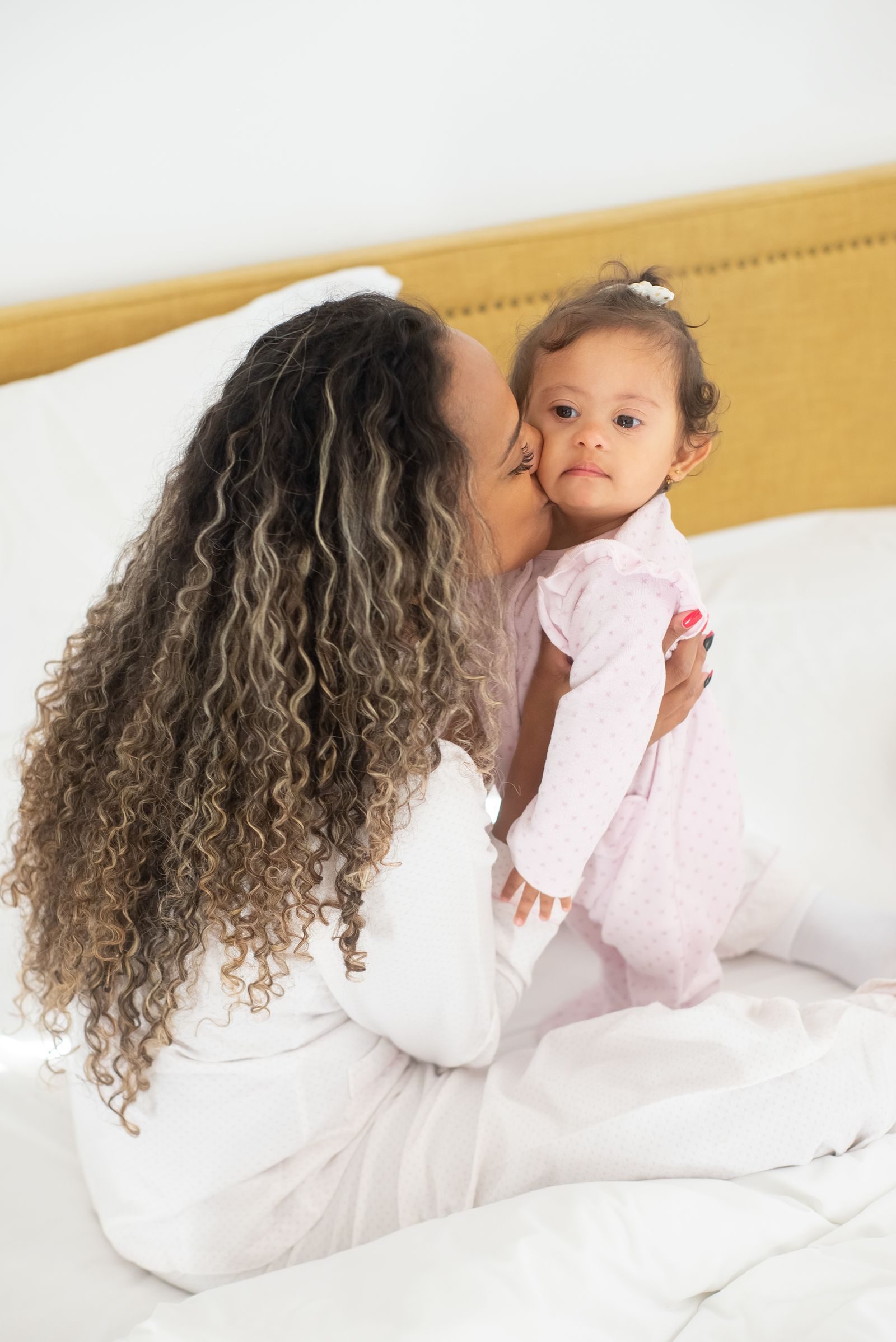 A loving mother gently kisses her baby while sitting on a cozy bed in a bright room, embracing affection and family bonds.
