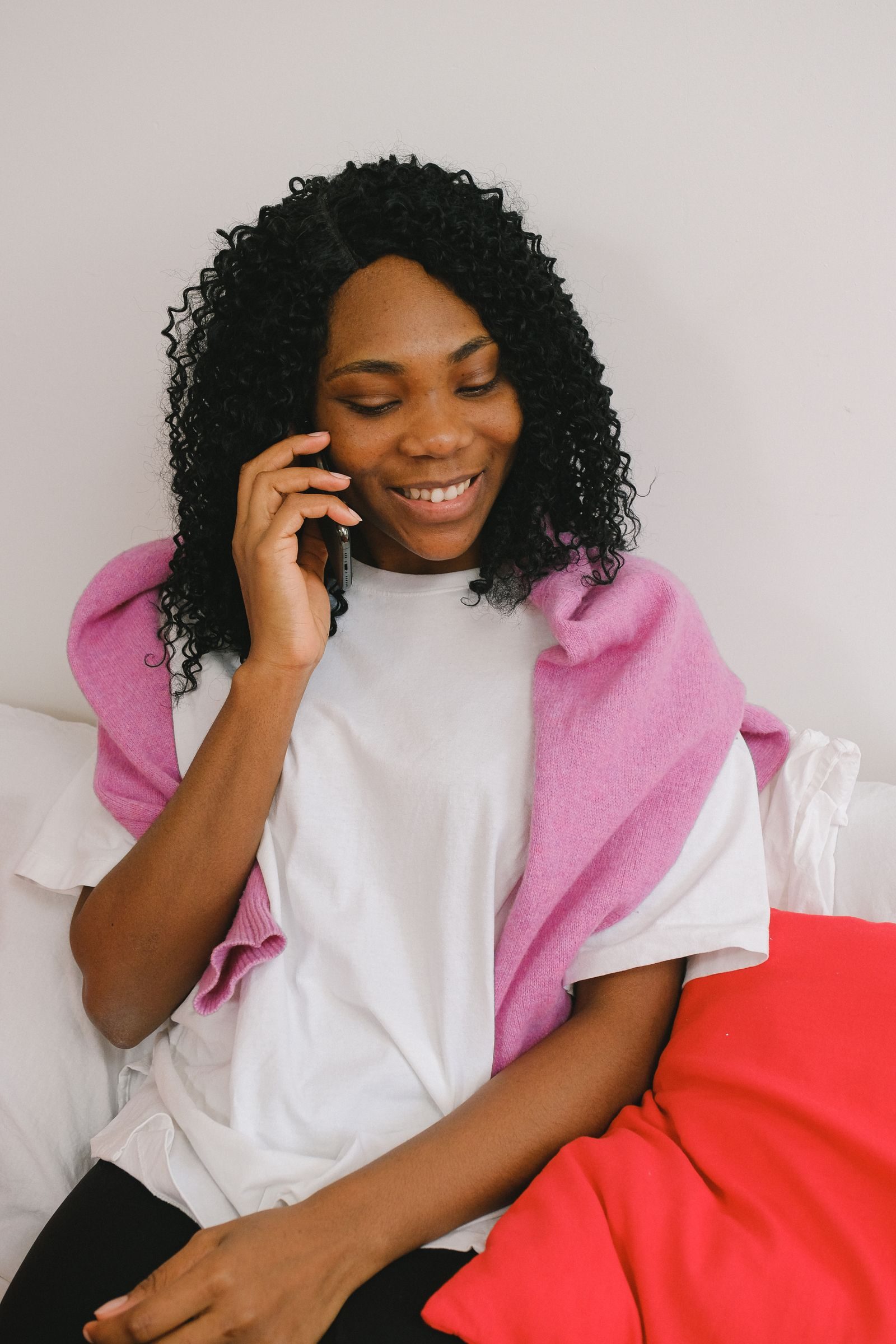 Content African American female having conversation on cellphone while sitting on comfortable bed with pillows in light bedroom at home