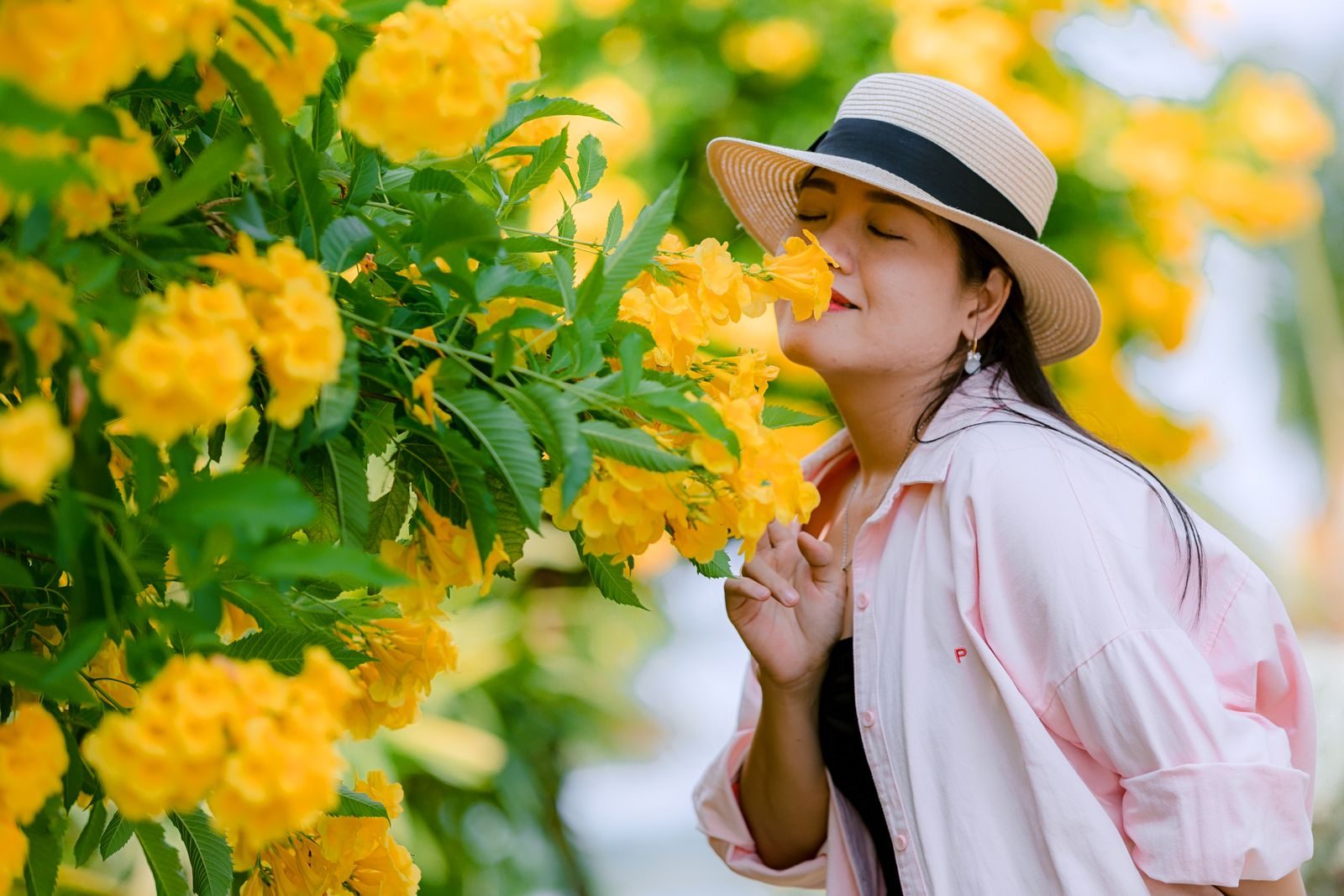 A woman in a hat joyfully breathes in the fragrance of vibrant yellow flowers outdoors.