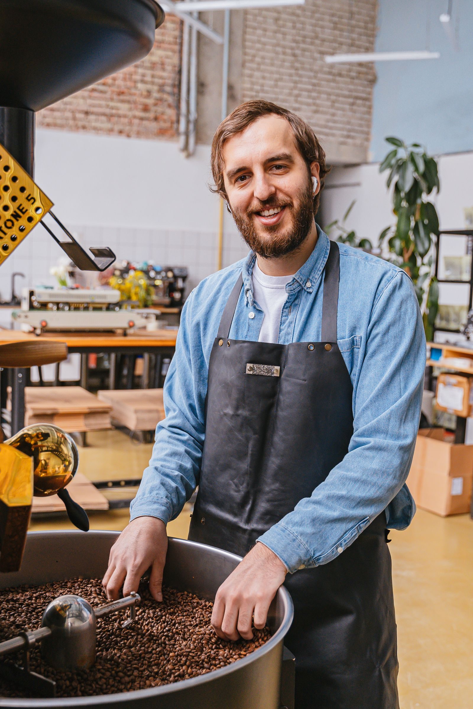 A male barista in a denim shirt and apron at a coffee shop roasting coffee beans.