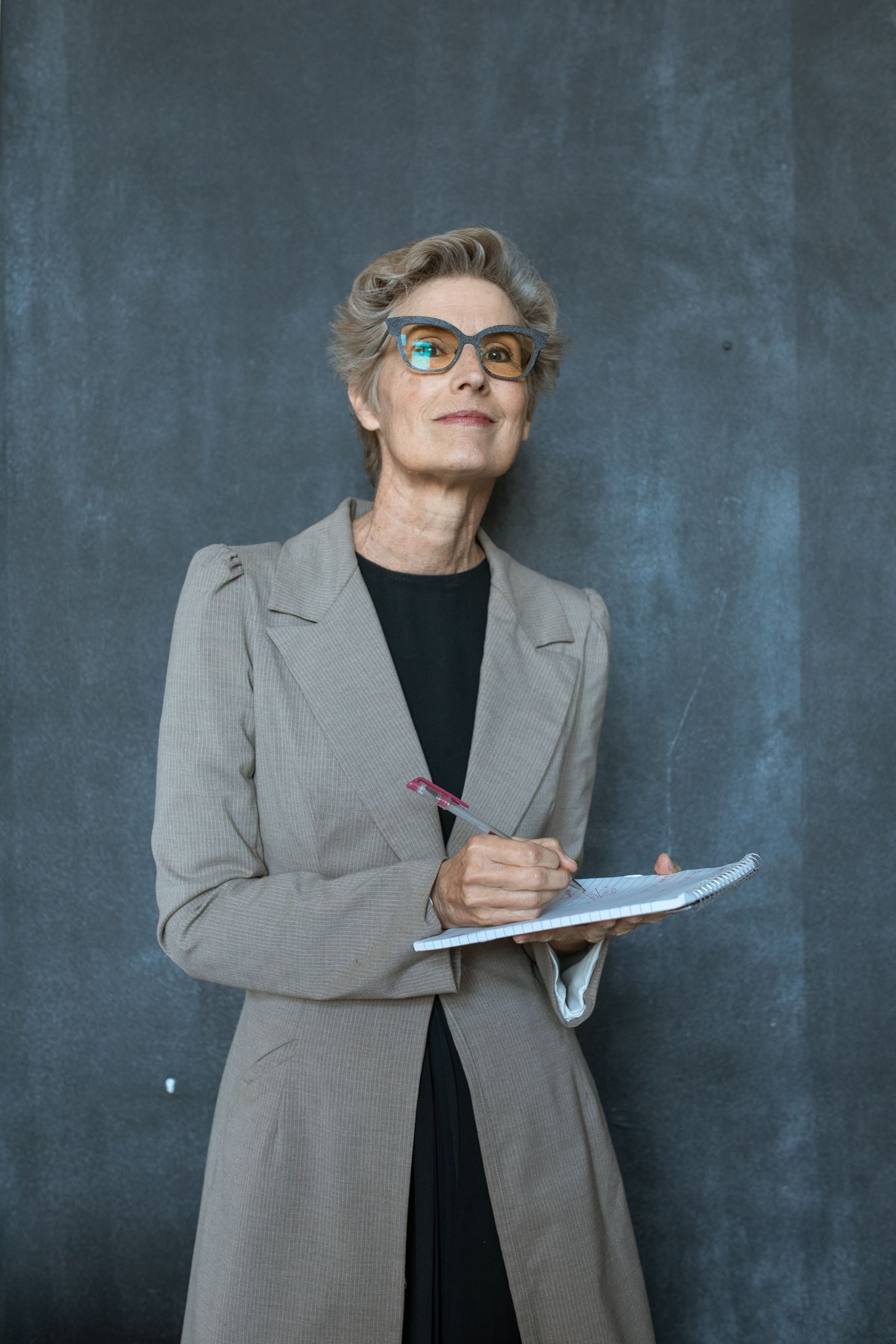 Sophisticated adult woman in glasses with gray coat, smiling while writing in a notebook.