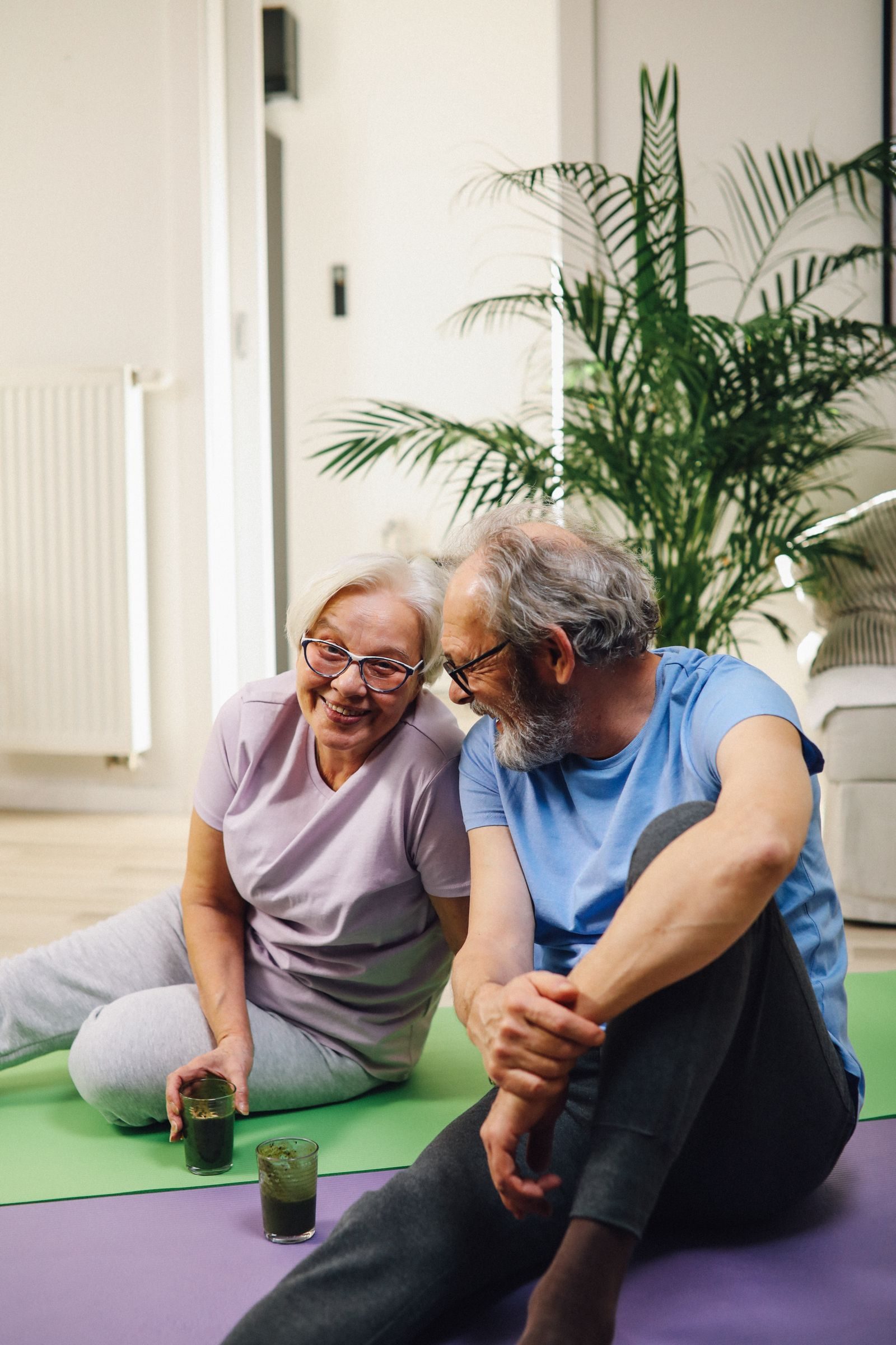 Elderly couple sitting on yoga mats, smiling with green smoothies, promoting healthy living.