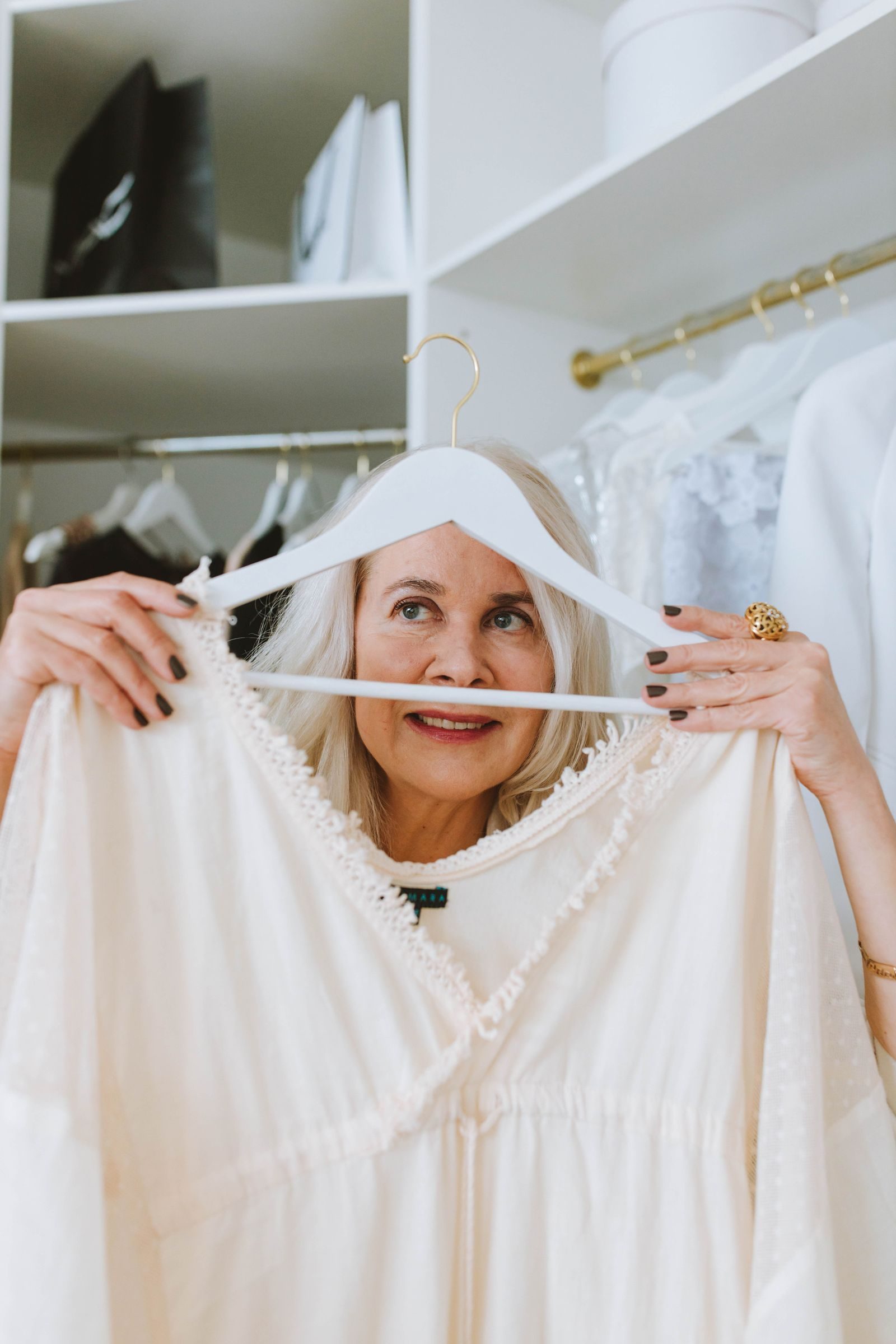 Elderly woman holding a white blouse in closet, smiling and browsing fashion options.