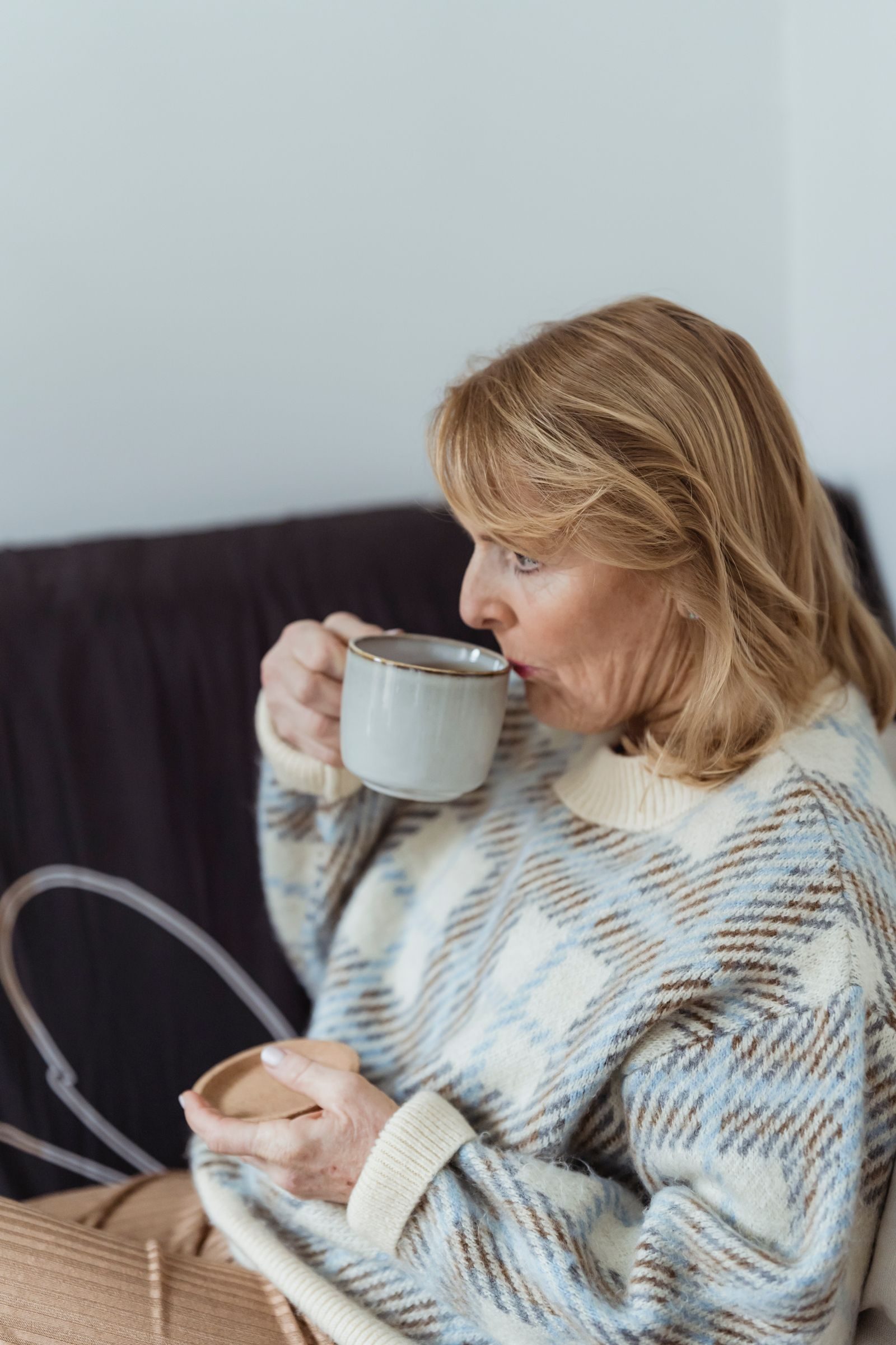 Side view of relaxed middle aged female with blond hair in warm sweater sitting on sofa and drinking hot aromatic coffee