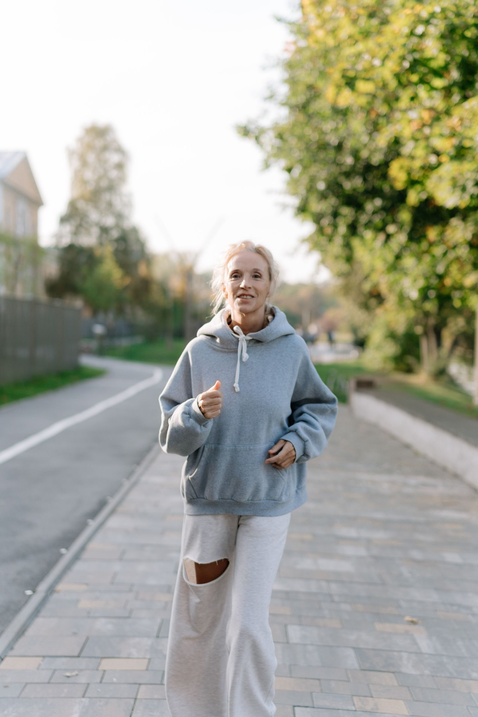Elderly woman jogging on a sidewalk in a green urban park area during the day.