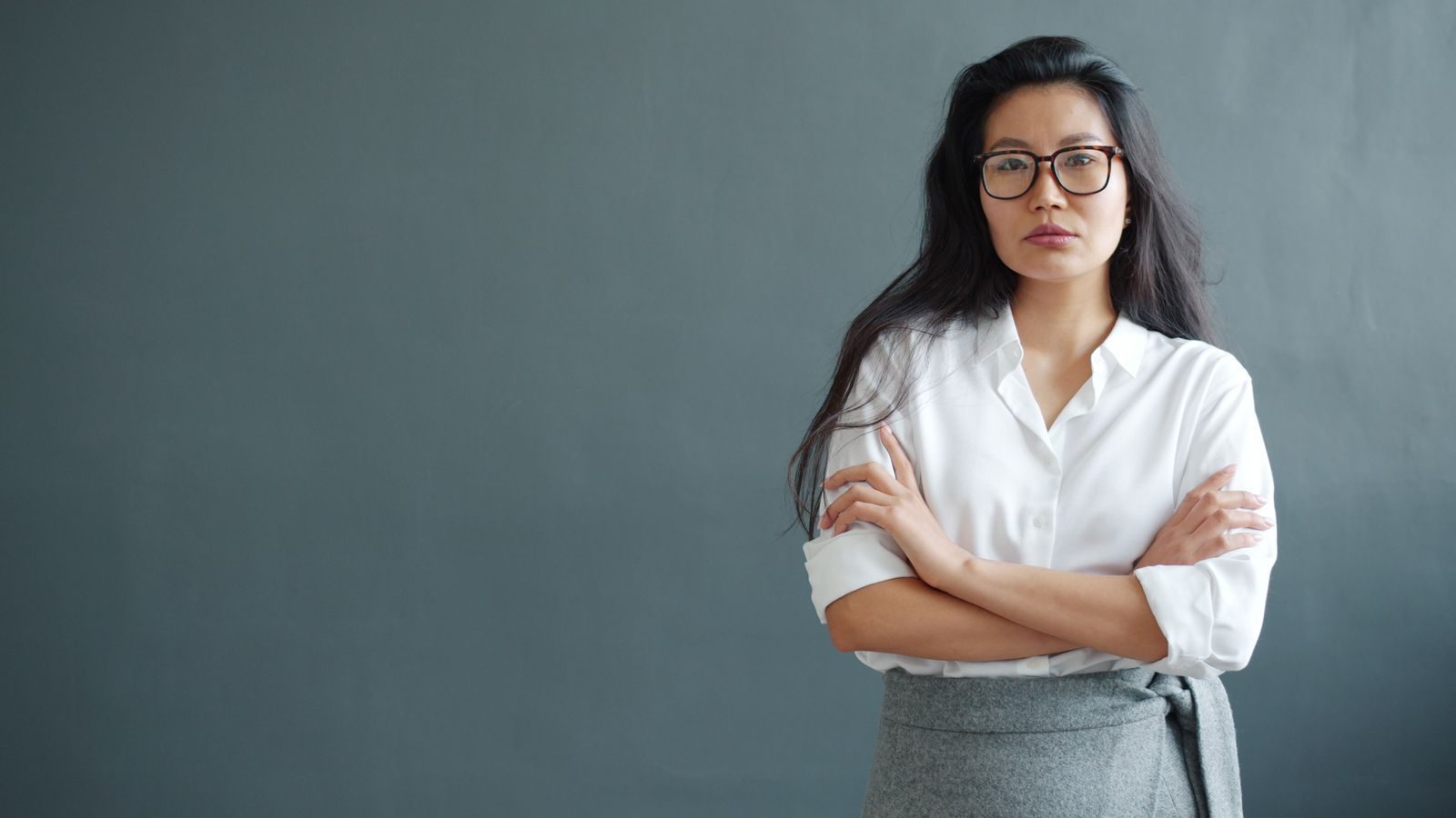 Confident woman in white shirt with arms crossed, wearing glasses, serious expression.