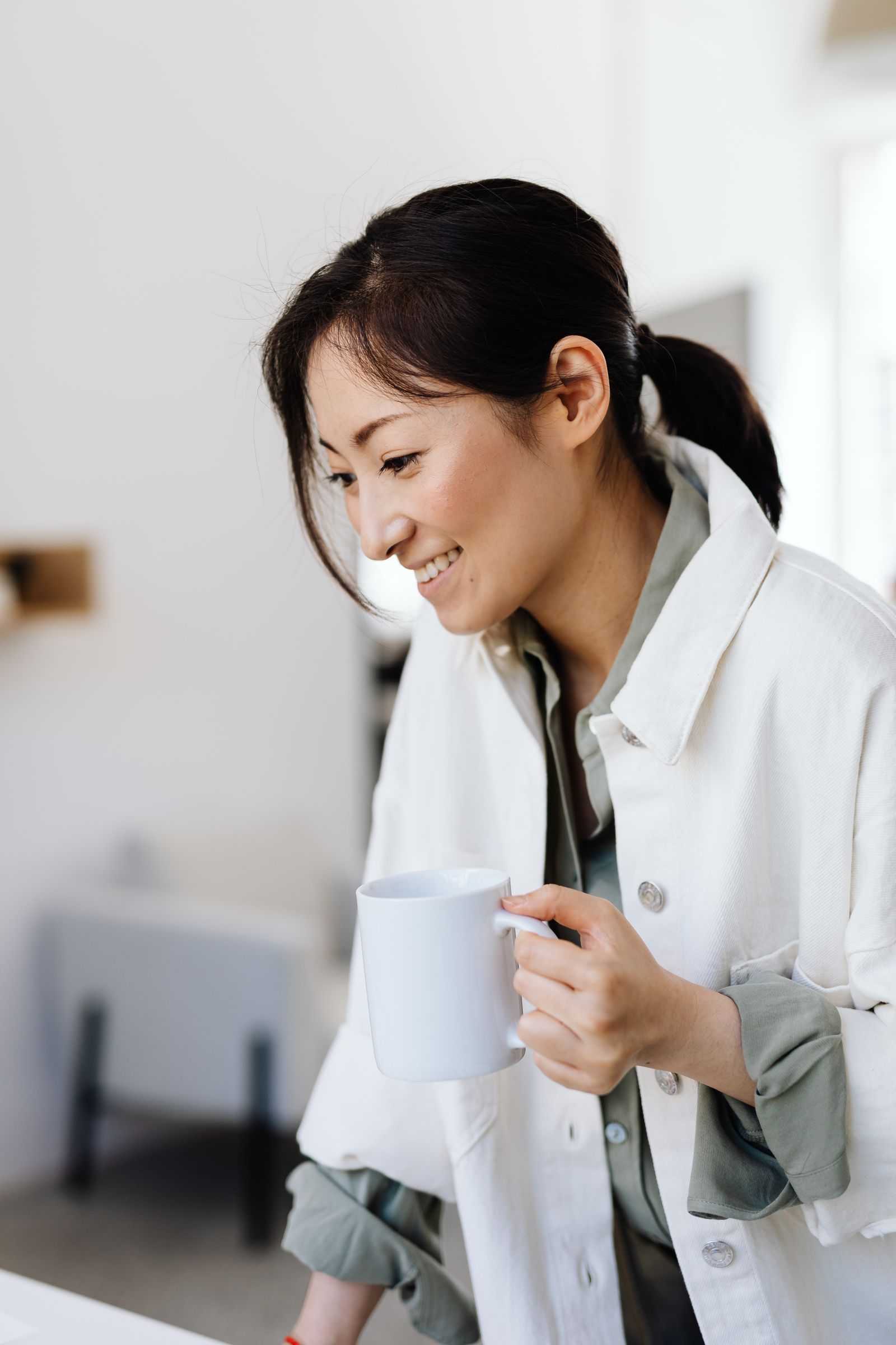Asian woman with ponytail smiling while holding a mug in a bright indoor setting, dressed casually.