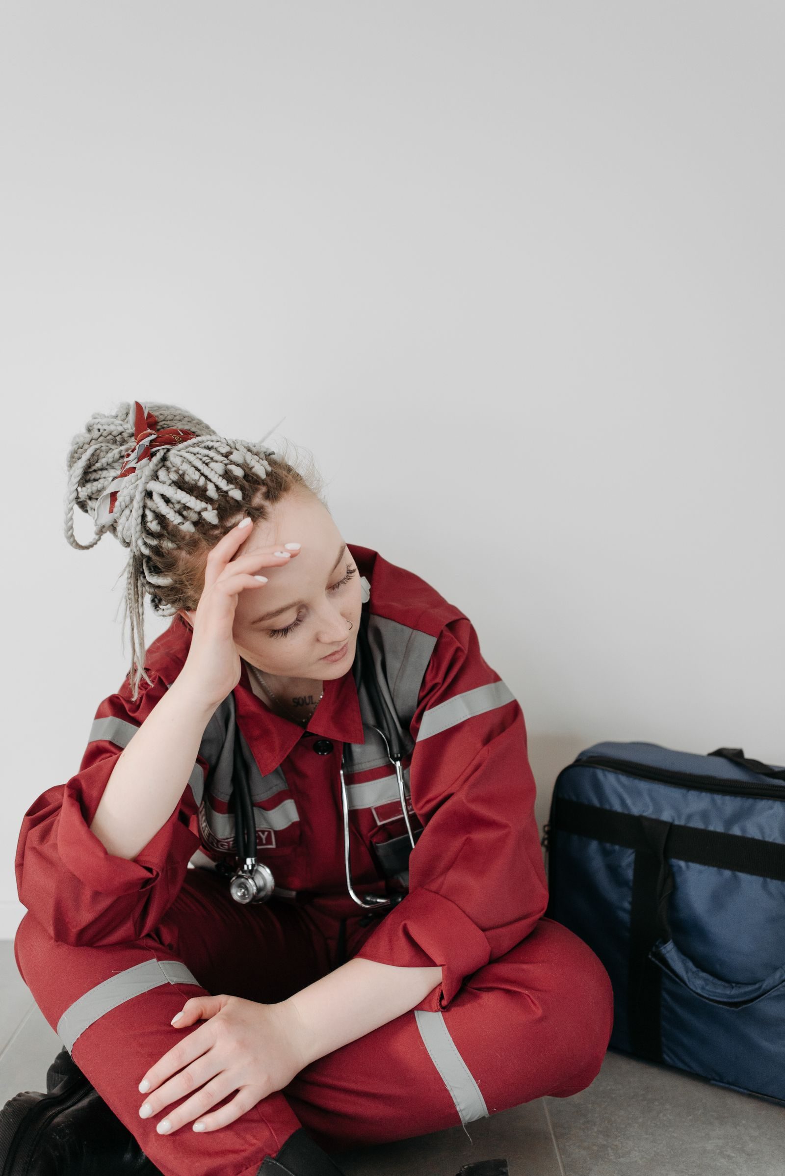 A tired female EMT in red uniform sitting on the floor, resting her head in her hand.