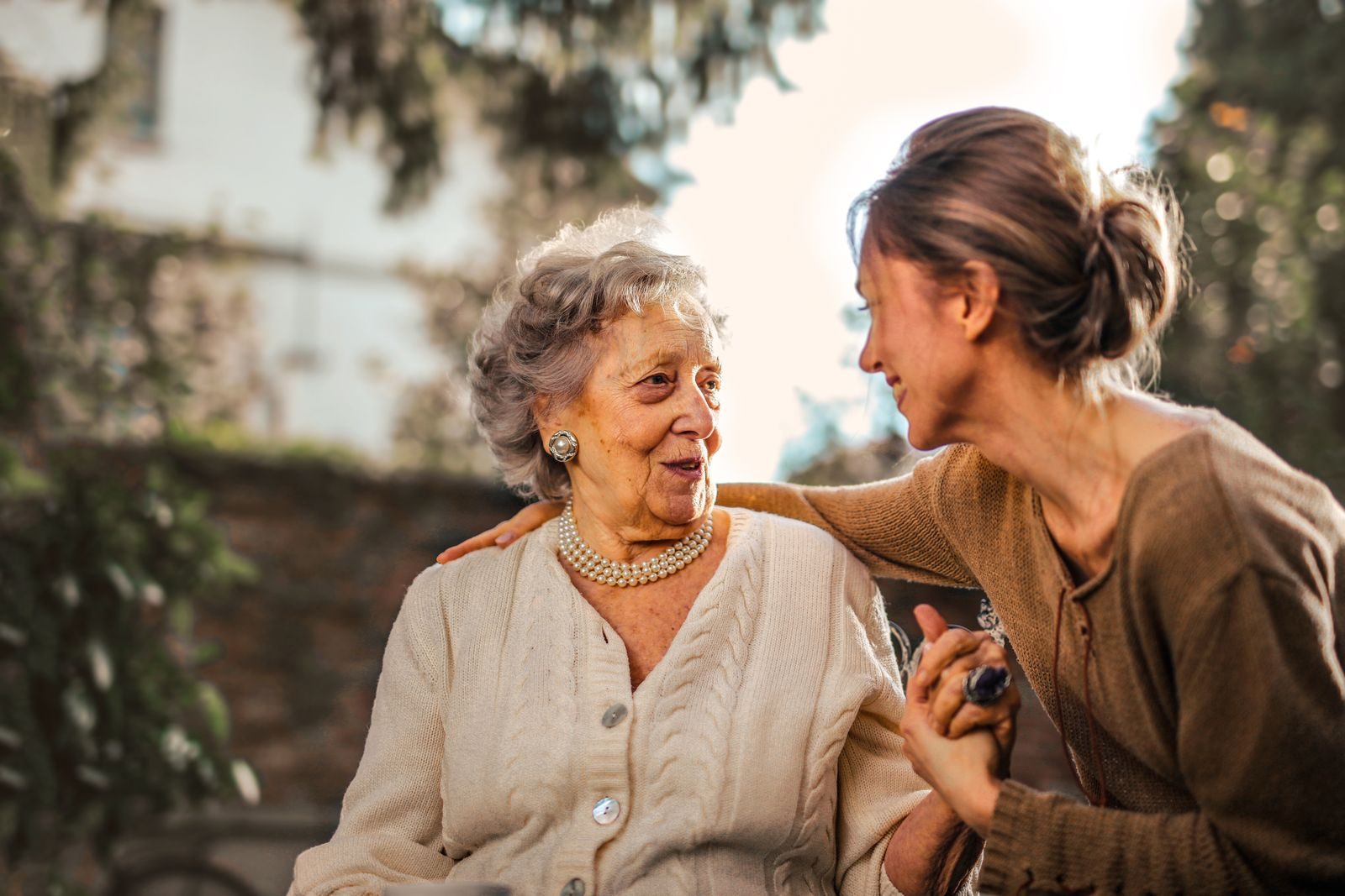 Low angle of cheerful aged lady and adult woman in casual clothes holding hands and smiling at each other while having meeting on yard against blurred green trees and facade of country house in suburb
