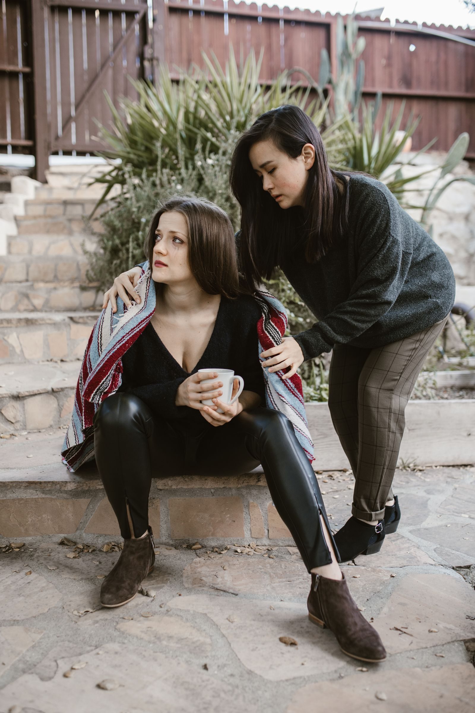 A woman comforts another on outdoor steps with a supportive gesture.