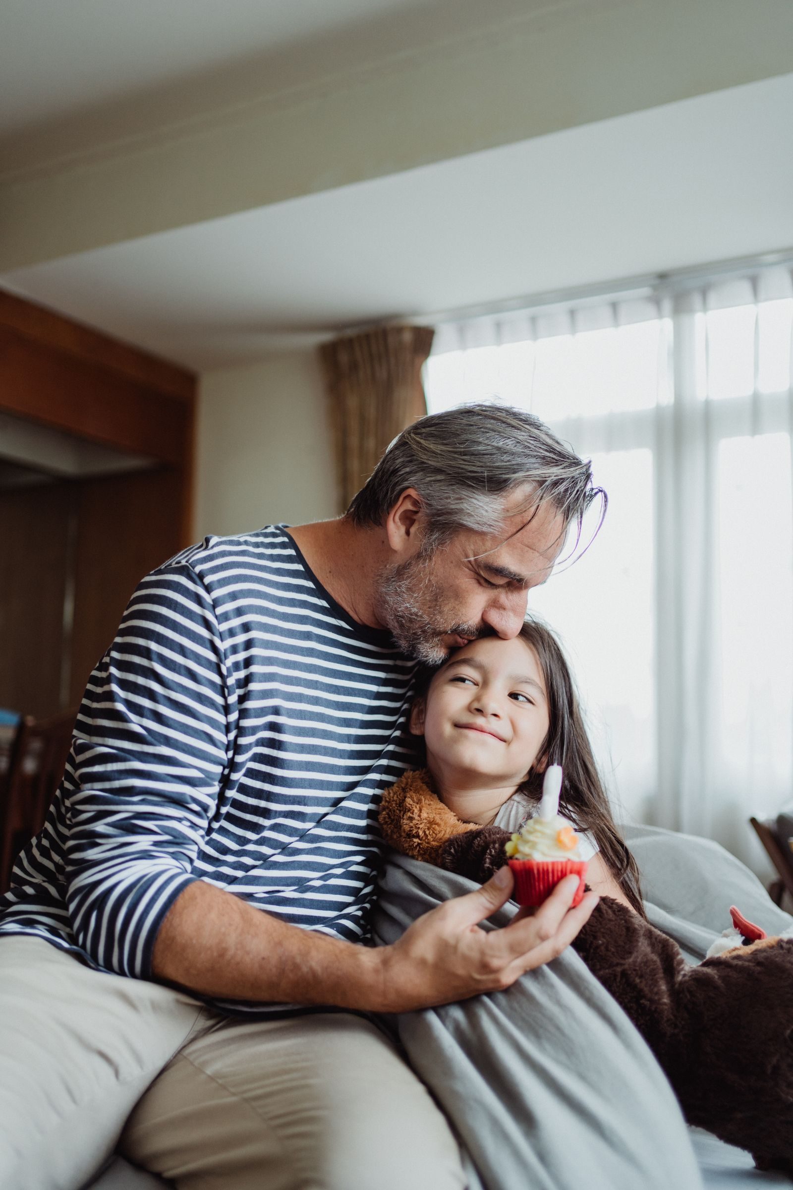 Adorable moment of a father kissing his daughter holding a cupcake indoors.
