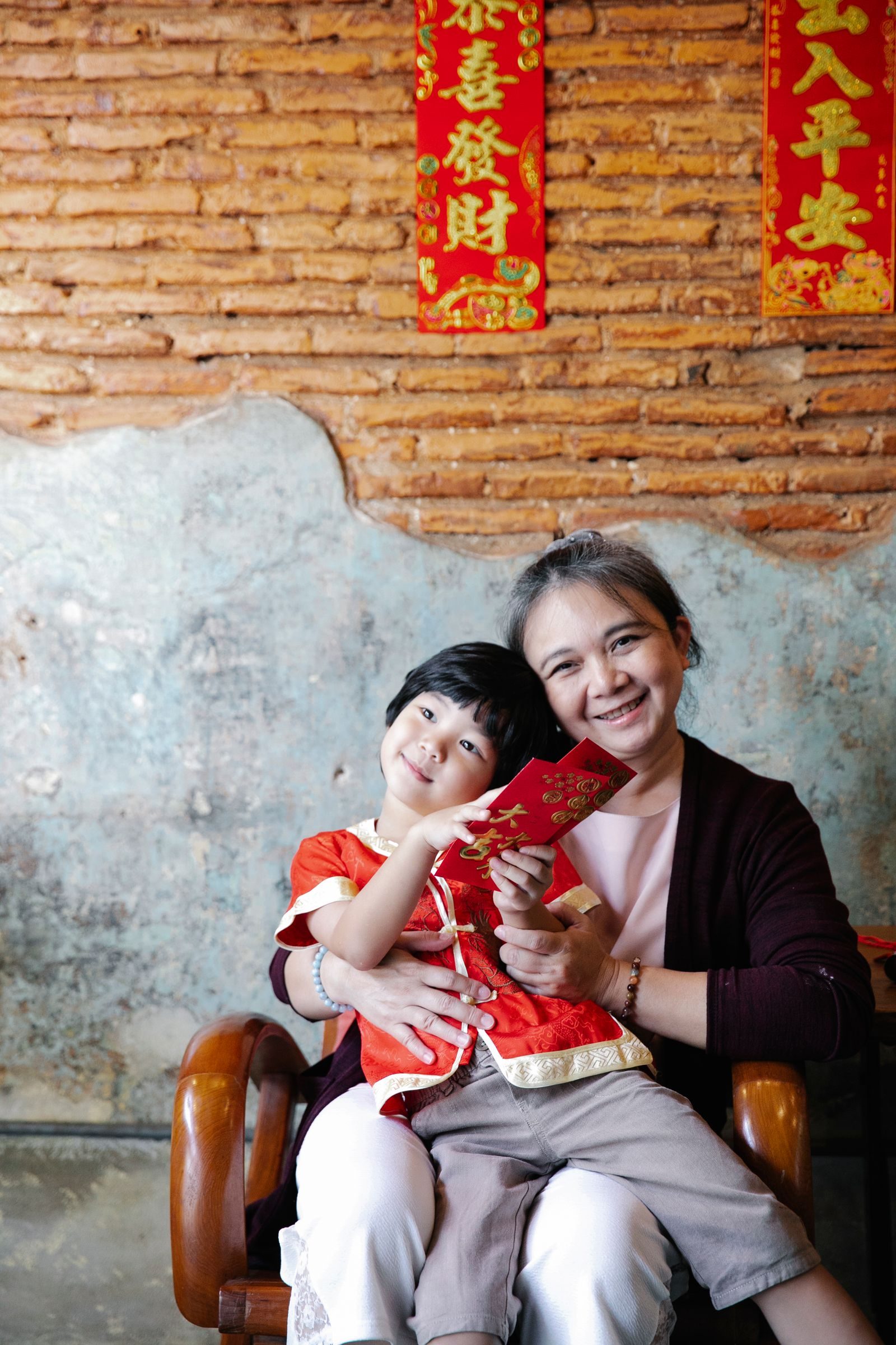 Positive senior ethnic woman smiling and looking at camera while hugging adorable little grandson holding traditional red envelopes during New Year holidays