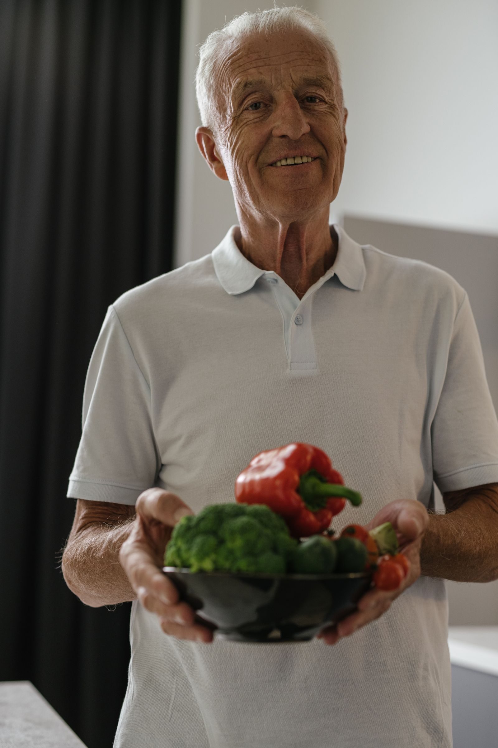 Elderly man happily holding a bowl of fresh vegetables indoors, promoting healthy lifestyle.