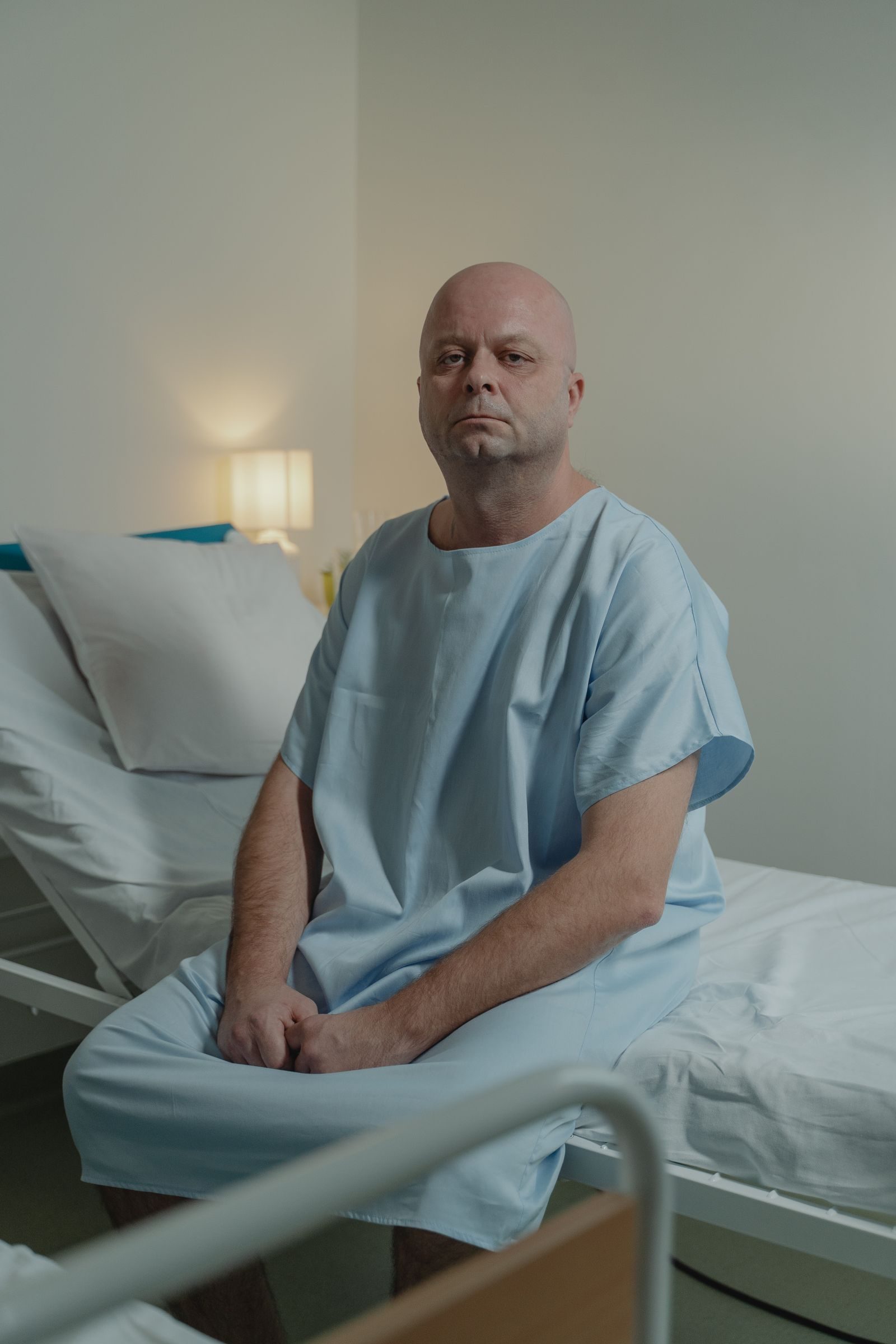 A bald man sitting on a hospital bed wearing a hospital gown, looking contemplative.