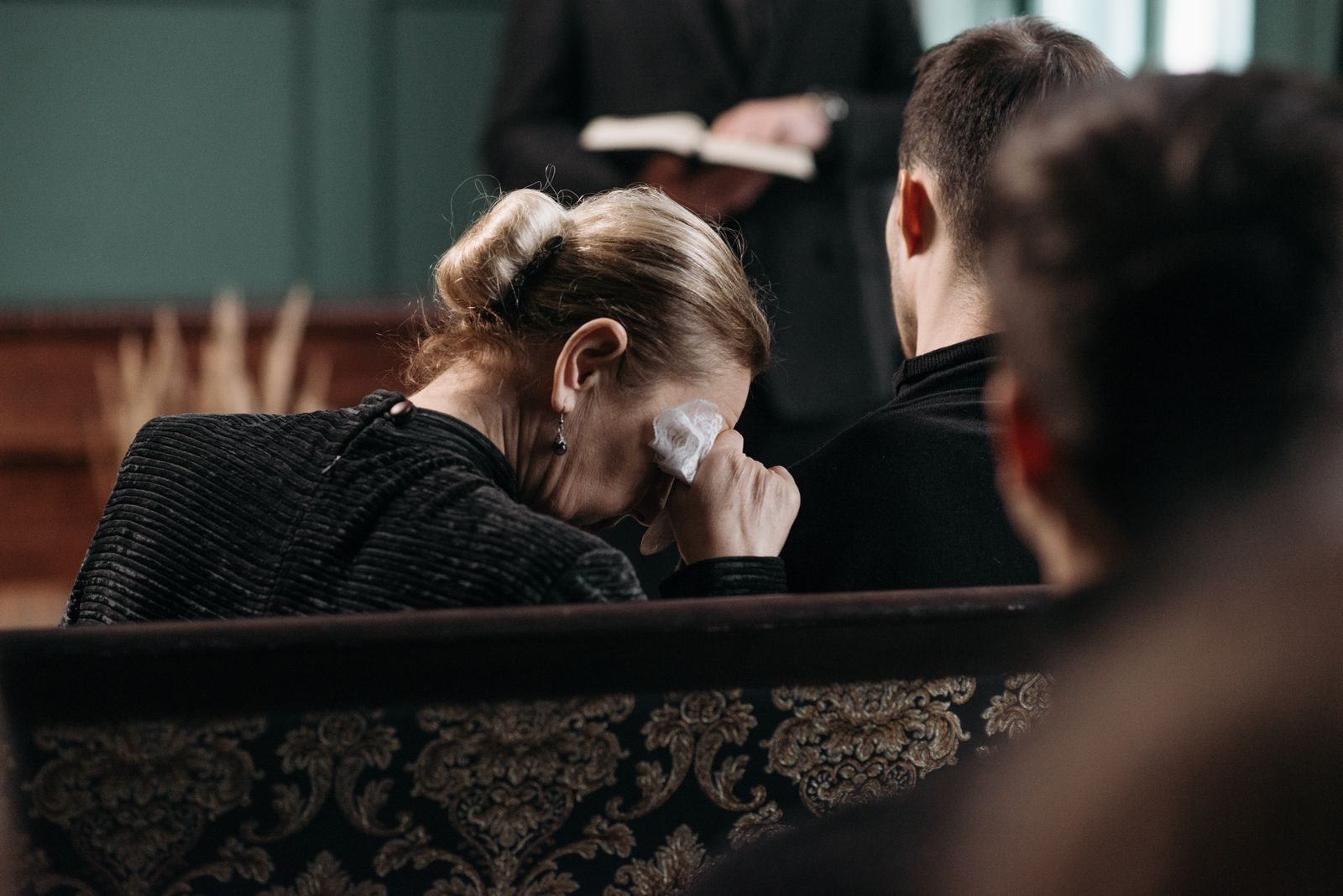 A grieving woman wipes tears during a heartfelt funeral ceremony in a somber indoor setting.