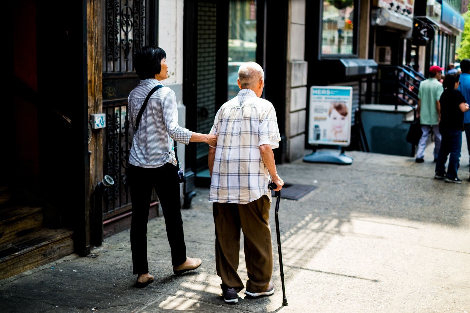 An elderly man with a cane is assisted by a younger companion walking along a busy New York City street.