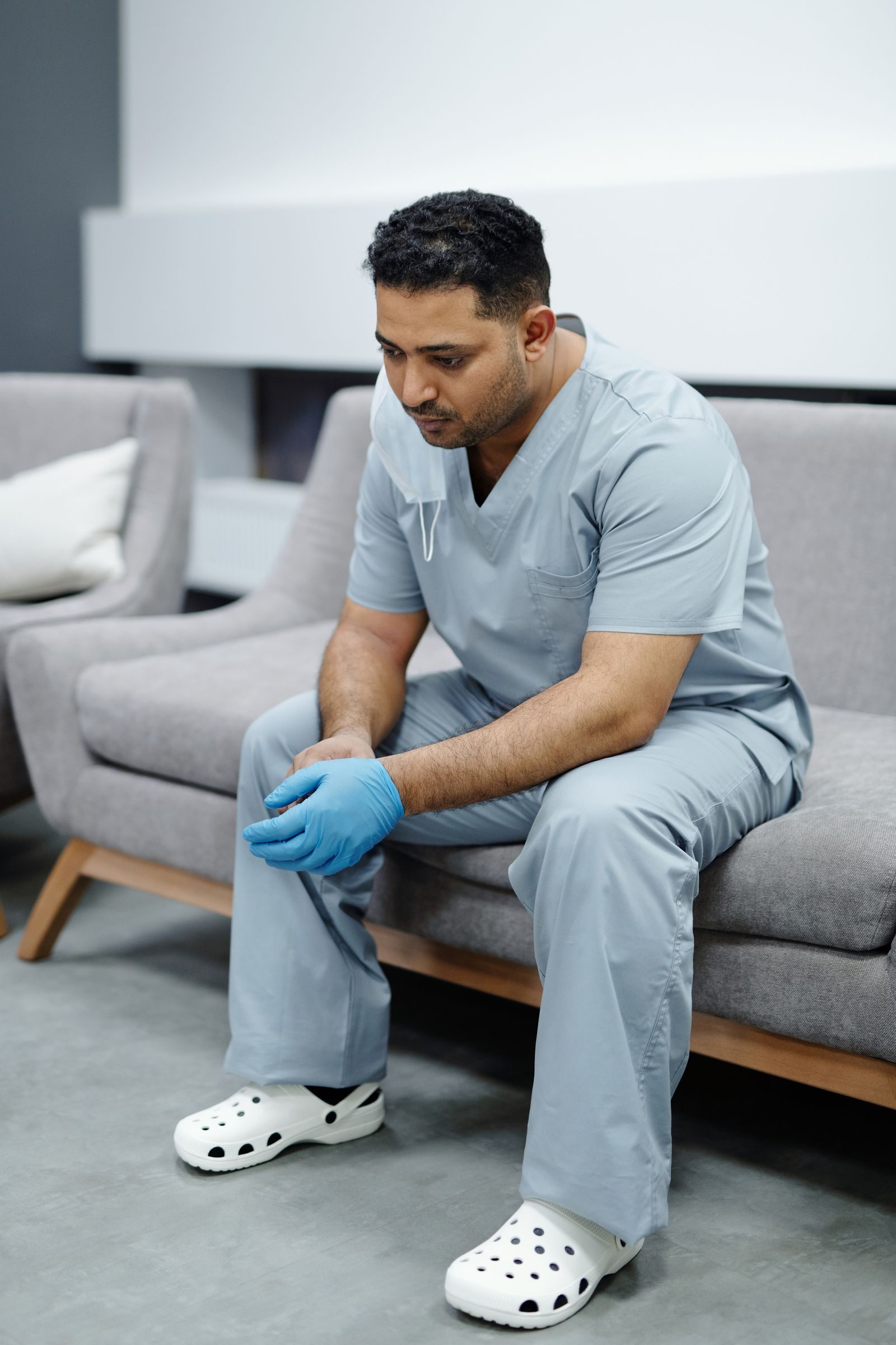 A tired healthcare professional in scrubs sits on a sofa, reflecting exhaustion after a long day.