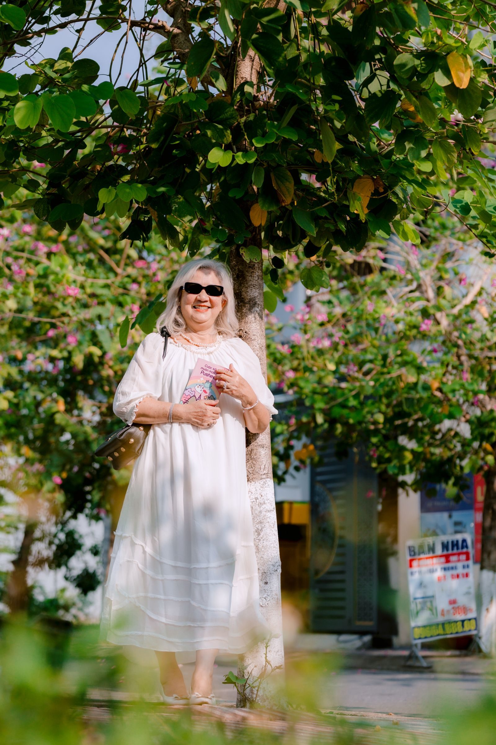 Elderly woman in a white dress, smiling and enjoying the sunny day under a tree.
