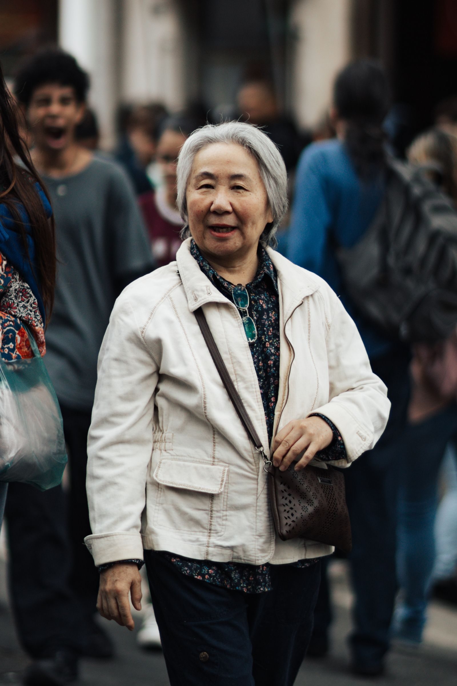 Smiling elderly woman with white hair walking in a bustling city crowd wearing a jacket.
