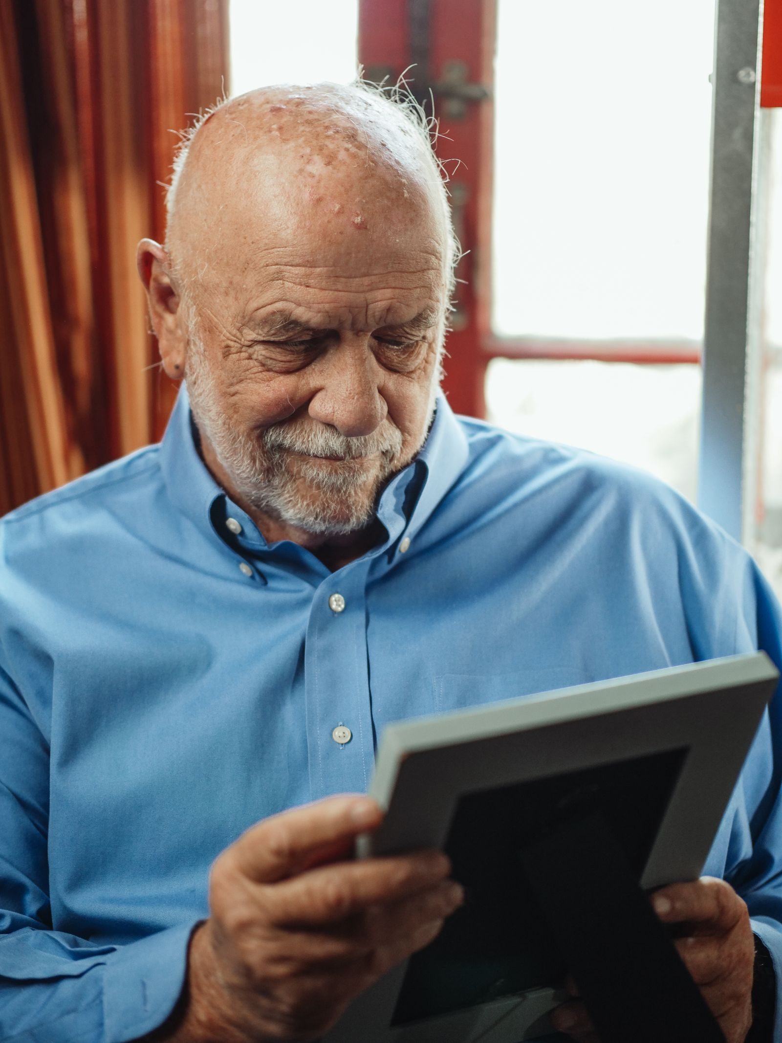 Senior man in blue shirt holding a photo frame, reflecting thoughtfully indoors.
