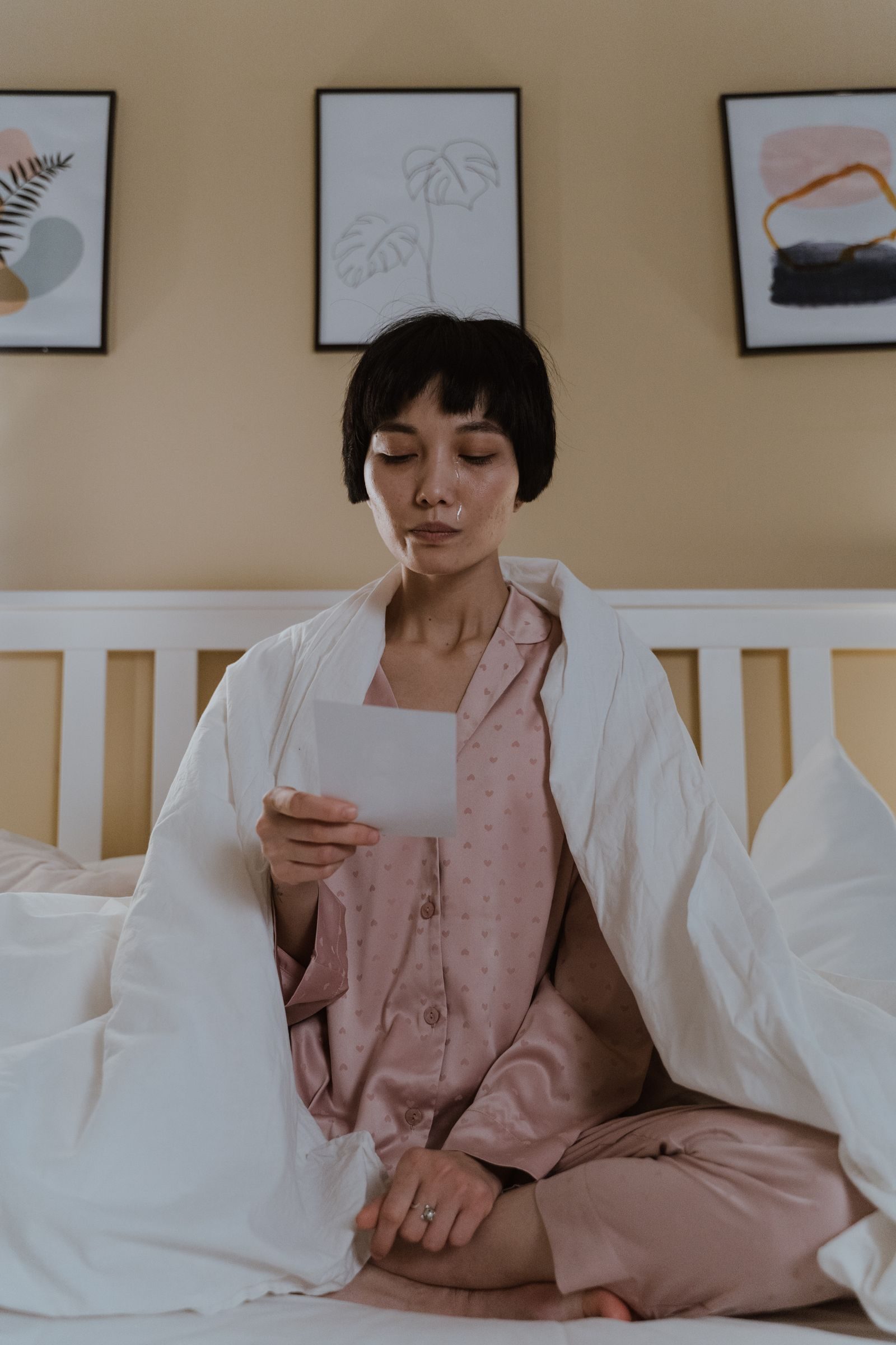 A woman in pajamas sits on a bed reading a letter, expressing emotion.