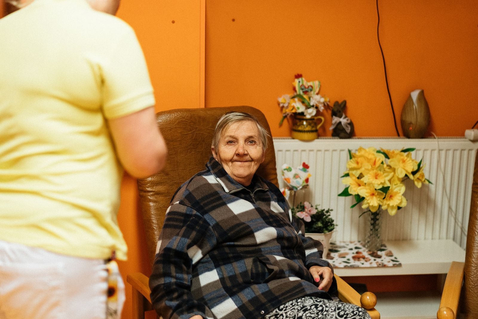 A warm scene of a senior woman sitting in an armchair at a retirement home with a caretaker nearby.