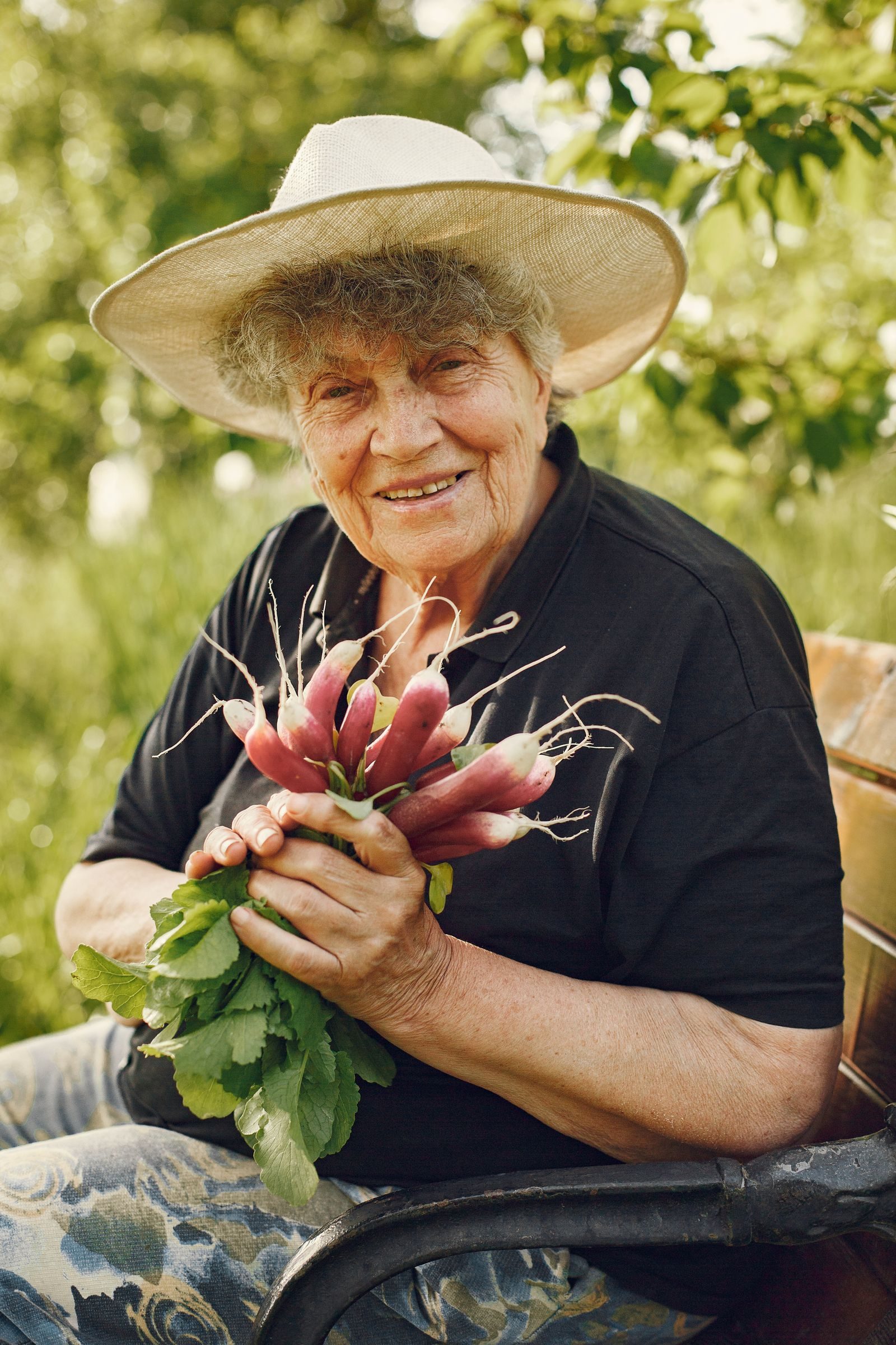 Elderly woman with a wide-brimmed hat holding fresh radishes in a sunny garden.