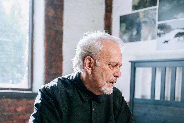 An elderly man in a black shirt sits pensively on a bed in a bright bedroom.