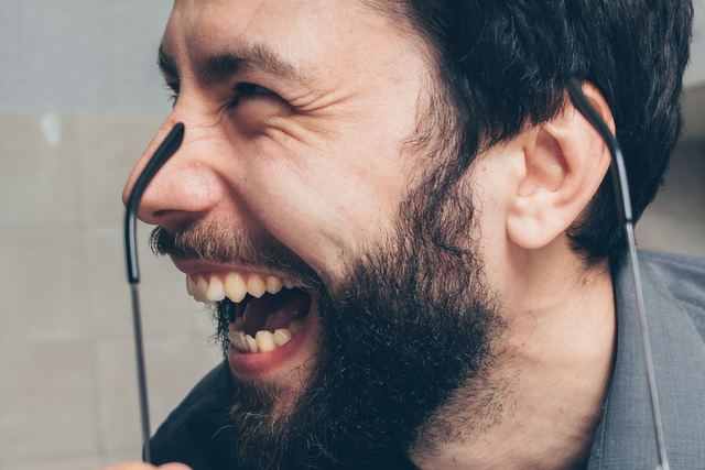 Close-up of a bearded man laughing joyfully, holding eyeglasses. Energetic and happy expression.