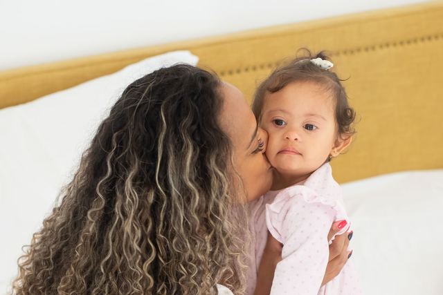 A loving mother gently kisses her baby while sitting on a cozy bed in a bright room, embracing affection and family bonds.