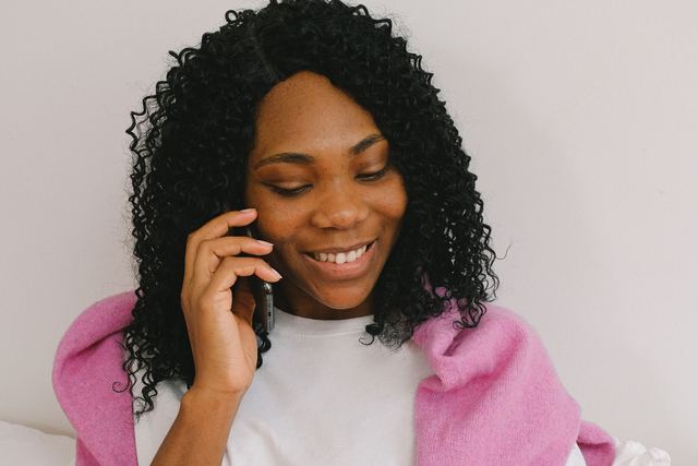 Content African American female having conversation on cellphone while sitting on comfortable bed with pillows in light bedroom at home