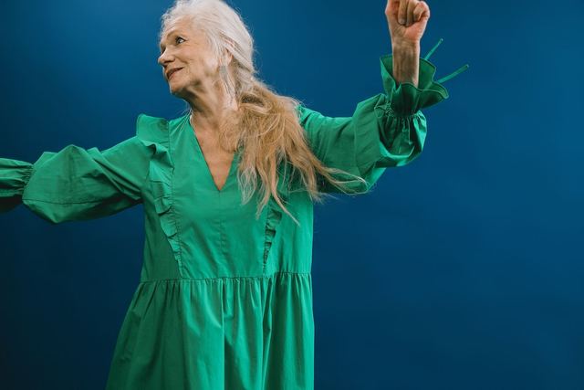 A joyful elderly woman in a green dress dances against a vibrant blue backdrop, showcasing timeless elegance.