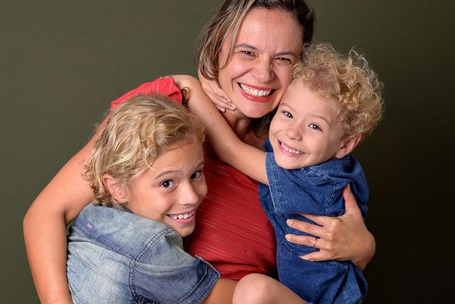A joyful mother embraces her smiling children against a green backdrop.