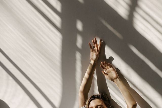 Smiling woman stretching with arms up in sunlight and shadows indoors.