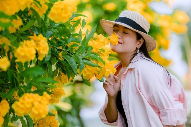 A woman in a hat joyfully breathes in the fragrance of vibrant yellow flowers outdoors.