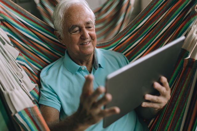 Smiling senior man uses a tablet while relaxing in a colorful hammock indoors.