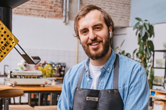A male barista in a denim shirt and apron at a coffee shop roasting coffee beans.