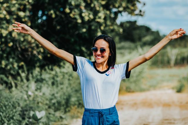 A smiling woman in sunglasses and a white shirt raises her arms in joy on a sunny day outdoors.