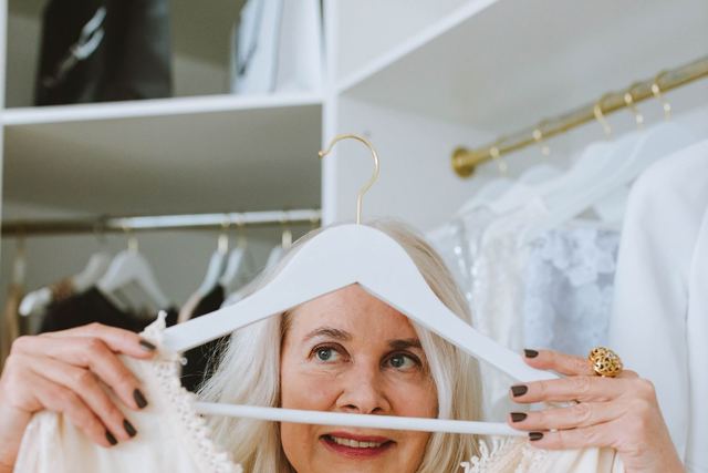 Elderly woman holding a white blouse in closet, smiling and browsing fashion options.