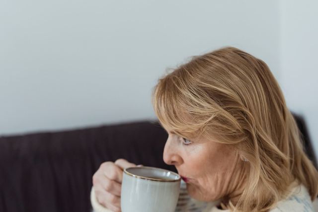 Side view of relaxed middle aged female with blond hair in warm sweater sitting on sofa and drinking hot aromatic coffee