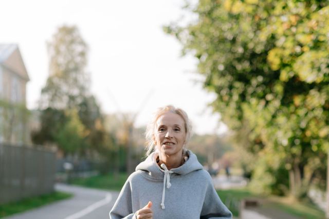 Elderly woman jogging on a sidewalk in a green urban park area during the day.