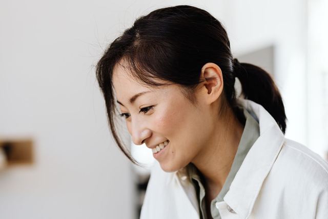 Asian woman with ponytail smiling while holding a mug in a bright indoor setting, dressed casually.