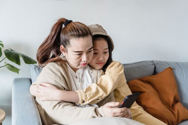 A heartwarming scene of a mother and daughter embracing on the sofa while looking at a smartphone.