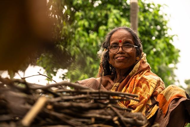 Elderly woman in sari smiling near stacked firewood, outdoors.