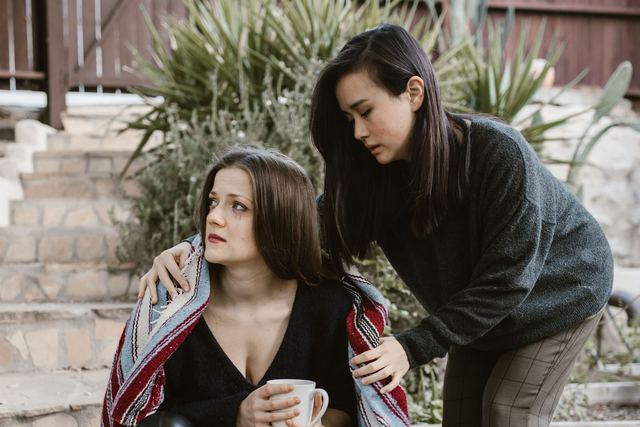 A woman comforts another on outdoor steps with a supportive gesture.