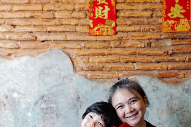 Positive senior ethnic woman smiling and looking at camera while hugging adorable little grandson holding traditional red envelopes during New Year holidays