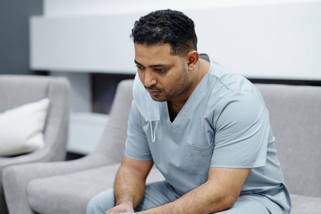 A tired healthcare professional in scrubs sits on a sofa, reflecting exhaustion after a long day.